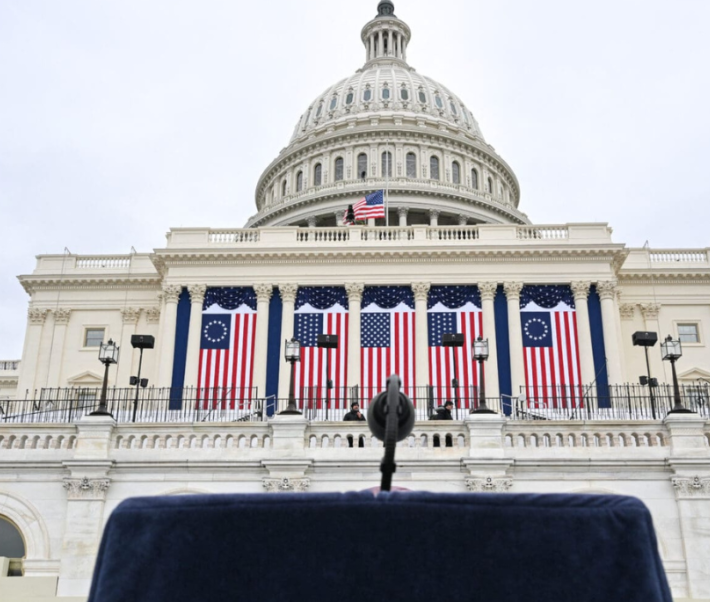 Photo du capitole aux États-Unis, avec un pupitre et un micro en premier plan et des drapeaux du pays en second plan.