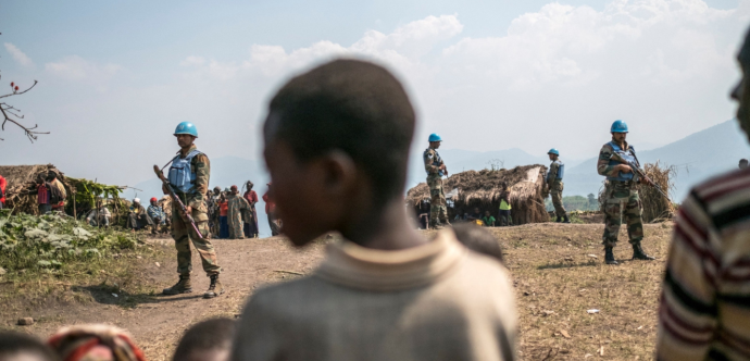 un groupe de soldats de la paix, armés et portant des casques et des gilets bleus, se tient devant un groupe de personnes déplacées.