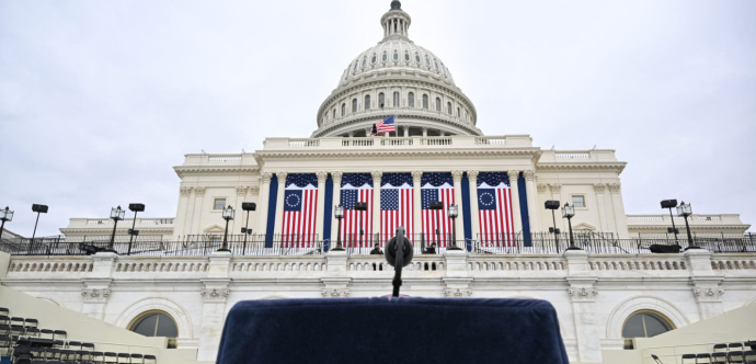 Photo du capitole aux États-Unis, avec un pupitre et un micro en premier plan et des drapeaux du pays en second plan.