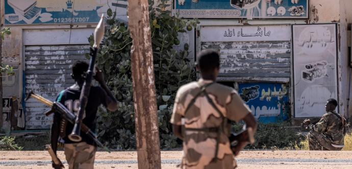 Trois hommes en tenue militaire et armés se trouvent dans une rue déserte. L’un porte un lance-roquettes sur l’épaule, un autre a une arme en bandoulière, et le troisième est assis au sol avec une mitrailleuse.