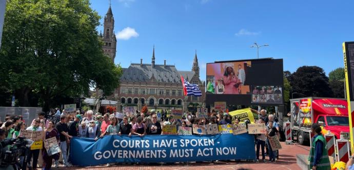Manifestation devant le Palais de la Paix à La Haye, aux Pays-Bas. Une grande bannière bleue portée par des manifestants affiche le message : “Courts have spoken – Governments must act now”. Les participants tiennent également d’autres pancartes sur le climat et la justice environnementale. Un écran géant diffuse le discours d'une intervenante, et des caméras de médias sont visibles au premier plan. L’événement se déroule sous un ciel bleu, dans une ambiance pacifique mais engagée.