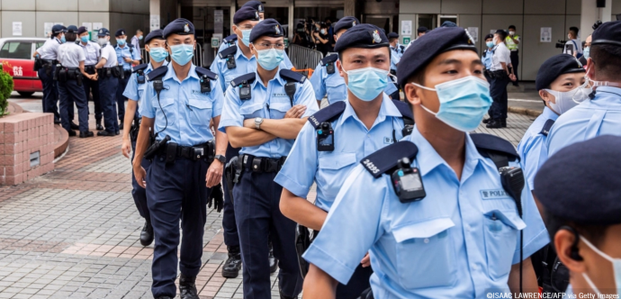 Groupe de policiers en uniforme bleu clair, portant des masques chirurgicaux et des bérets, marchant en file dans un espace urbain pavé. Certains portent des caméras corporelles et des équipements à la ceinture. À l’arrière-plan, d’autres agents discutent entre eux près d’un bâtiment officiel. 