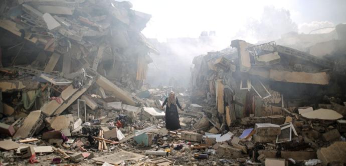 Une femme marche seule au milieu des décombres d’immeubles effondrés, entourée de gravats, de poutres brisées et de fragments de béton. Une fumée épaisse s’élève à l’arrière-plan, suggérant une récente explosion ou attaque. L’atmosphère est lourde, marquée par la destruction et le chaos.