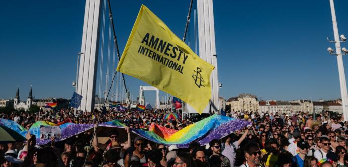 Une foule nombreuse participe à une marche ou manifestation sur un pont, portant des drapeaux arc-en-ciel et un grand drapeau jaune d’Amnesty International. Les participants sourient, brandissent des pancartes et des bannières multicolores sous un ciel bleu clair. L’événement semble festif et engagé, promouvant les droits humains et la cause LGBTQIA+.
