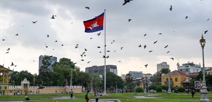 « Place publique à Phnom Penh, Cambodge, avec un grand mât portant le drapeau cambodgien flottant au vent. De nombreux oiseaux volent dans le ciel nuageux. Des passants se promènent sur les pelouses bien entretenues, entourées de bâtiments coloniaux colorés, d’arbres et de lampadaires dorés. En arrière-plan, on aperçoit des immeubles modernes.