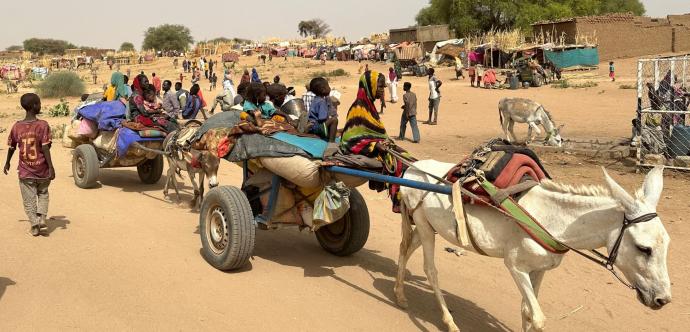 Scène de vie dans un camp de réfugiés en milieu désertique. Des ânes tirent des charrettes chargées de sacs, d'effets personnels et de plusieurs enfants assis, certains enveloppés dans des couvertures colorées. Des adultes marchent à côté, et en arrière-plan, on voit de nombreuses personnes, des abris de fortune, des huttes et des arbres. Le sol est sableux, et l'atmosphère suggère chaleur et aridité. 