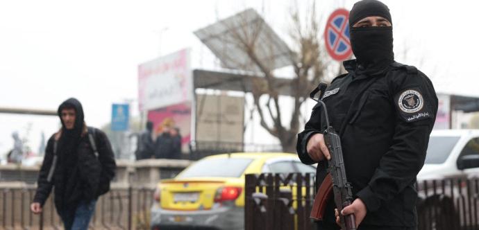 « Un homme en uniforme noir, masqué et tenant un fusil d’assaut, se tient en position de garde dans une rue. À l’arrière-plan, un piéton en capuche marche devant des voitures, dont un taxi jaune, et des panneaux urbains, par temps couvert. 