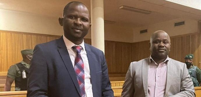 « Deux hommes en costume se tiennent debout dans un tribunal, derrière une barrière en bois. L’un porte une cravate à motifs rouges et bleus, tandis que l’autre est en chemise à carreaux et veste grise. En arrière-plan, deux gardes en uniforme vert surveillent la salle d’audience.