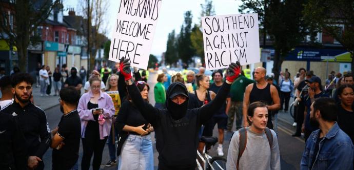 Manifestation dans une rue avec un groupe de personnes. Au centre, une personne masquée vêtue de noir tient deux pancartes en l'air indiquant : "Migrants welcome here" et "Southend against racism". D'autres manifestants sont visibles en arrière-plan