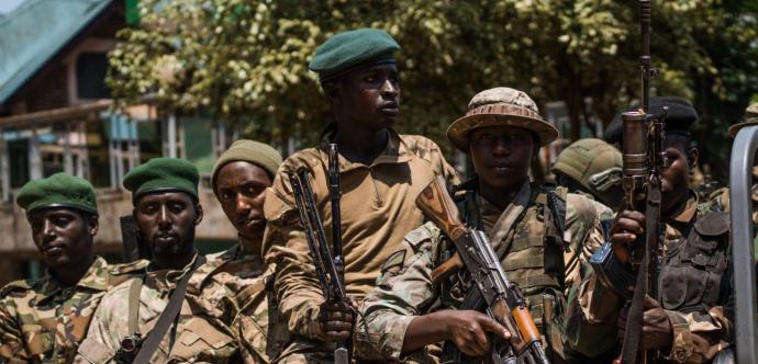 Groupe de soldats armés portant des uniformes militaires et des bérets verts ou noirs. Certains tiennent des fusils d’assaut, tandis qu’ils se tiennent serrés les uns contre les autres, semblant prêts à une opération. En arrière-plan, on aperçoit des arbres et un bâtiment.