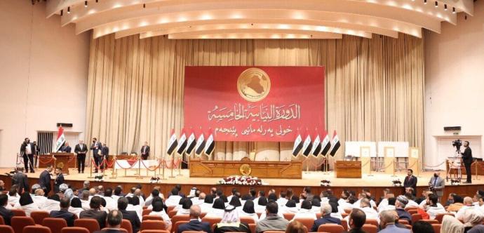 « Vue d’ensemble d’une séance parlementaire dans un grand hémicycle en Irak. Des parlementaires sont assis face à une estrade ornée de multiples drapeaux irakiens et d’un grand panneau rouge portant des inscriptions en arabe et un emblème doré représentant la carte du pays. Plusieurs responsables se tiennent debout sur la scène, tandis que des caméras et agents de sécurité sont présents dans la salle. 