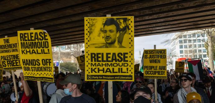 Manifestation sous un pont avec des pancartes jaunes portant des slogans en anglais tels que « Release Mahmoud Khalil ! », « Hands off our students ! » et « Resistance is justified when people are occupied ! ». Une foule de personnes, certaines portant un keffieh ou un masque, brandit des pancartes et un drapeau palestinien, exprimant leur solidarité avec la Palestine.