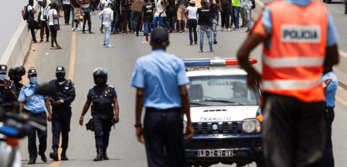 « Manifestation sur une route, avec un groupe de personnes rassemblées en arrière-plan. Au premier plan, plusieurs policiers, certains portant des casques et des gilets, encadrent la scène, tandis qu'une voiture de police marquée "Trânsito" est visible. Un policier en gilet fluorescent observe la situation