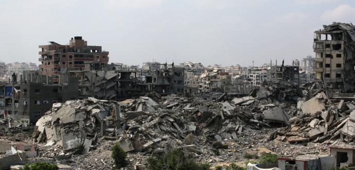 Vue panoramique d’une ville fortement détruite, avec des immeubles effondrés et des tas de gravats. Au premier plan, quelques arbres et petites structures encore debout contrastent avec les ruines massives en arrière-plan. Le ciel est nuageux, accentuant l’atmosphère de désolation