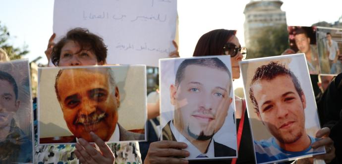 Un groupe de personnes manifeste en tenant des photos de proches disparus. Trois portraits d’hommes souriants ou sérieux sont clairement visibles, protégés par du plastique. À l’arrière-plan, d’autres participants tiennent des pancartes avec du texte en arabe. La scène se déroule en extérieur, en plein jour.