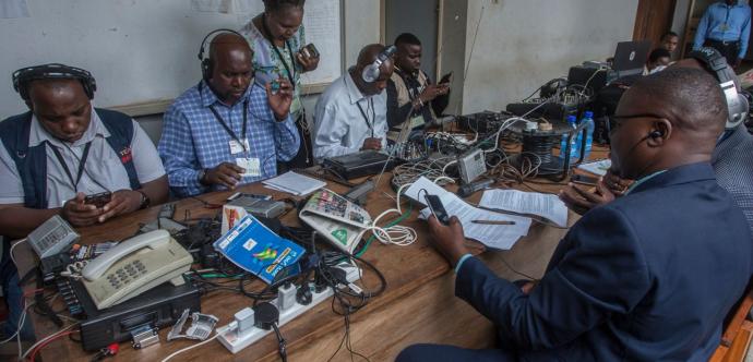 Un groupe de journalistes et techniciens assis autour d’une grande table en bois, équipée de téléphones fixes, câbles, ordinateurs portables, enregistreurs et autres matériels audio. Tous portent des casques sur les oreilles et consultent des téléphones ou des documents, concentrés sur leur travail. L’ambiance reflète l’intensité d’une salle de presse en pleine activité.