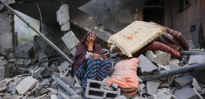 Une femme assise au milieu des décombres d’un bâtiment détruit, la tête dans la main en signe de détresse. Autour d’elle, des blocs de béton brisés, des poutres métalliques tordues et quelques matelas recouverts de poussière témoignent de la destruction causée par le bombardement.