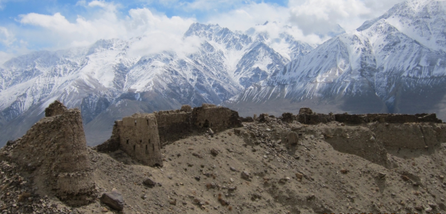 L'image montre un paysage accidenté et époustouflant d'un village pamiri niché dans les montagnes du Pamir, au Tadjikistan. Les ruines d'anciennes structures en pierre sont visibles au premier plan, perchées sur un terrain sec et rocailleux. À l'arrière-plan, les imposants pics enneigés de la chaîne du Pamir s'élèvent de façon spectaculaire dans un ciel bleu partiellement nuageux.