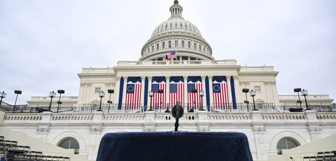 Photo du capitole aux États-Unis, avec un pupitre et un micro en premier plan et des drapeaux du pays en second plan.