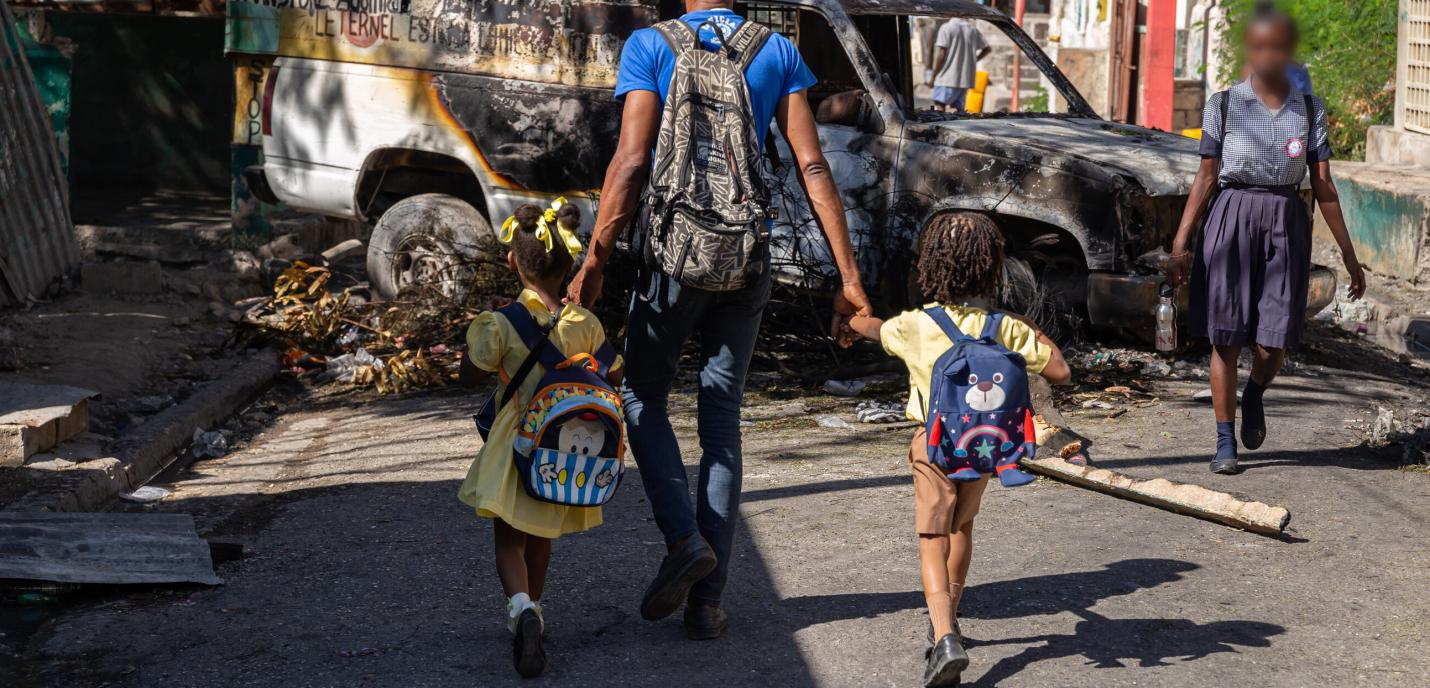 Un père emmène ses deux enfants à l'école, tenant leur main. Devant eux, une voiture calcinée bloque la rue.