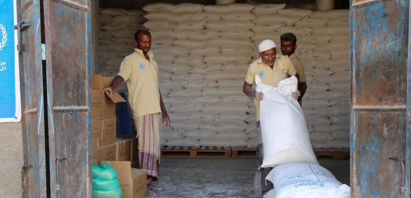 Trois hommes travaillant pour le Programme alimentaire mondial (PAM), dont l’un soulève un sac de riz. En arrière-plan, plusieurs sacs de riz empilés.