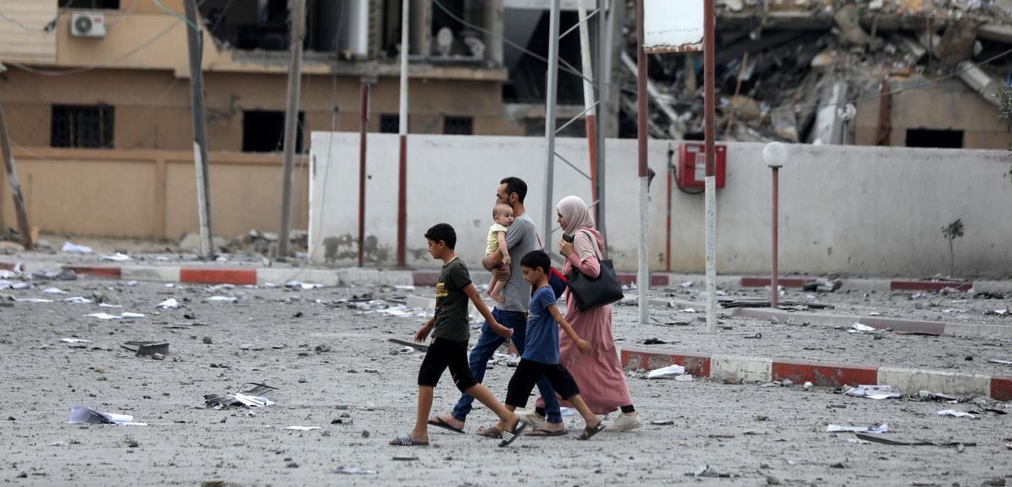 Une famille composée de deux adultes et quatre enfants traverse une rue jonchée de débris et de papiers, dans une zone fortement endommagée par des destructions. En arrière-plan, des bâtiments partiellement effondrés témoignent de récentes violences. Le père porte un bébé dans les bras, tandis que les autres enfants marchent à côté de leur mère, qui est voilée. L’atmosphère est tendue, marquée par les traces visibles d’un conflit.
