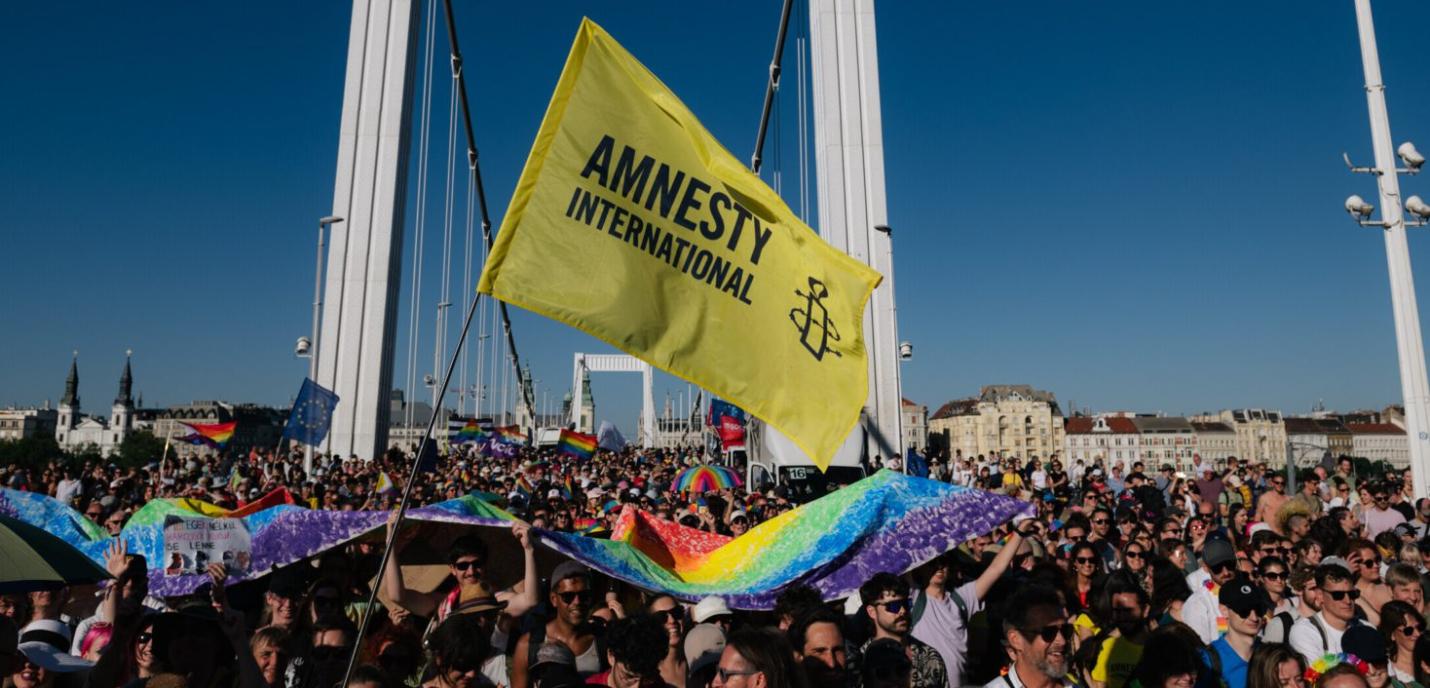 Une foule nombreuse participe à une marche ou manifestation sur un pont, portant des drapeaux arc-en-ciel et un grand drapeau jaune d’Amnesty International. Les participants sourient, brandissent des pancartes et des bannières multicolores sous un ciel bleu clair. L’événement semble festif et engagé, promouvant les droits humains et la cause LGBTQIA+.