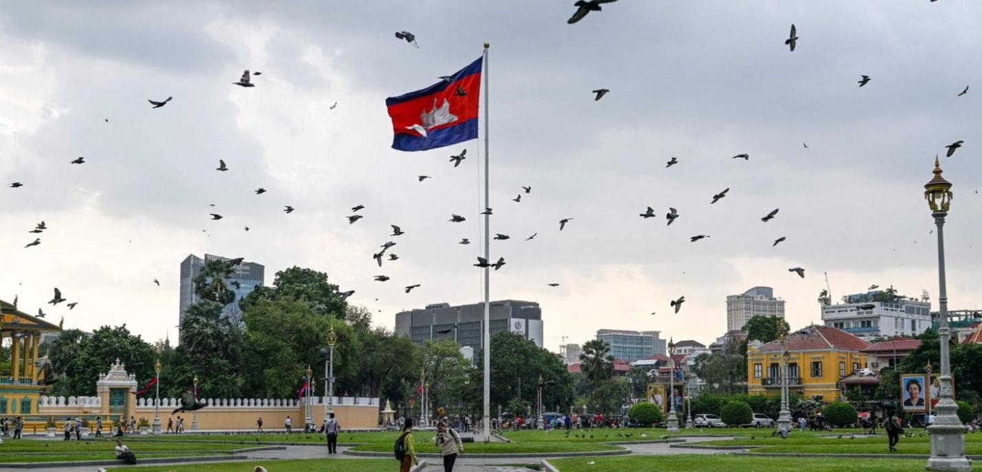 « Place publique à Phnom Penh, Cambodge, avec un grand mât portant le drapeau cambodgien flottant au vent. De nombreux oiseaux volent dans le ciel nuageux. Des passants se promènent sur les pelouses bien entretenues, entourées de bâtiments coloniaux colorés, d’arbres et de lampadaires dorés. En arrière-plan, on aperçoit des immeubles modernes.