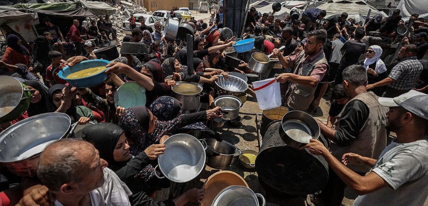 Des bénévoles distribuent de la nourriture à une foule dense de personnes, femmes, hommes et enfants, tendant des casseroles, bols et seaux dans une zone urbaine détruite. L’image reflète une situation d’urgence humanitaire, avec des bâtiments en ruine et des signes de conflit à l’arrière-plan.