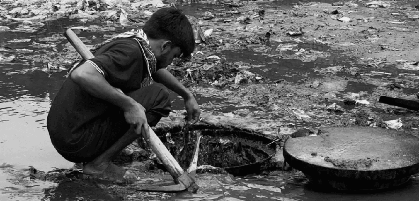 Un homme accroupi travaille dans des eaux sales et boueuses, manipulant un outil semblable à une pioche près d’une bouche d’égout ouverte. L’environnement est jonché de détritus et reflète des conditions de travail dangereuses et insalubres.