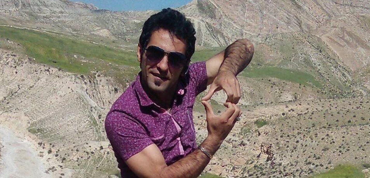 « Un homme portant une chemise violette à motifs et des lunettes de soleil pose en plein air, devant un paysage montagneux aride et vallonné. Il forme un geste avec ses mains comme s’il encadrait quelque chose entre ses doigts, en souriant légèrement à la caméra. 