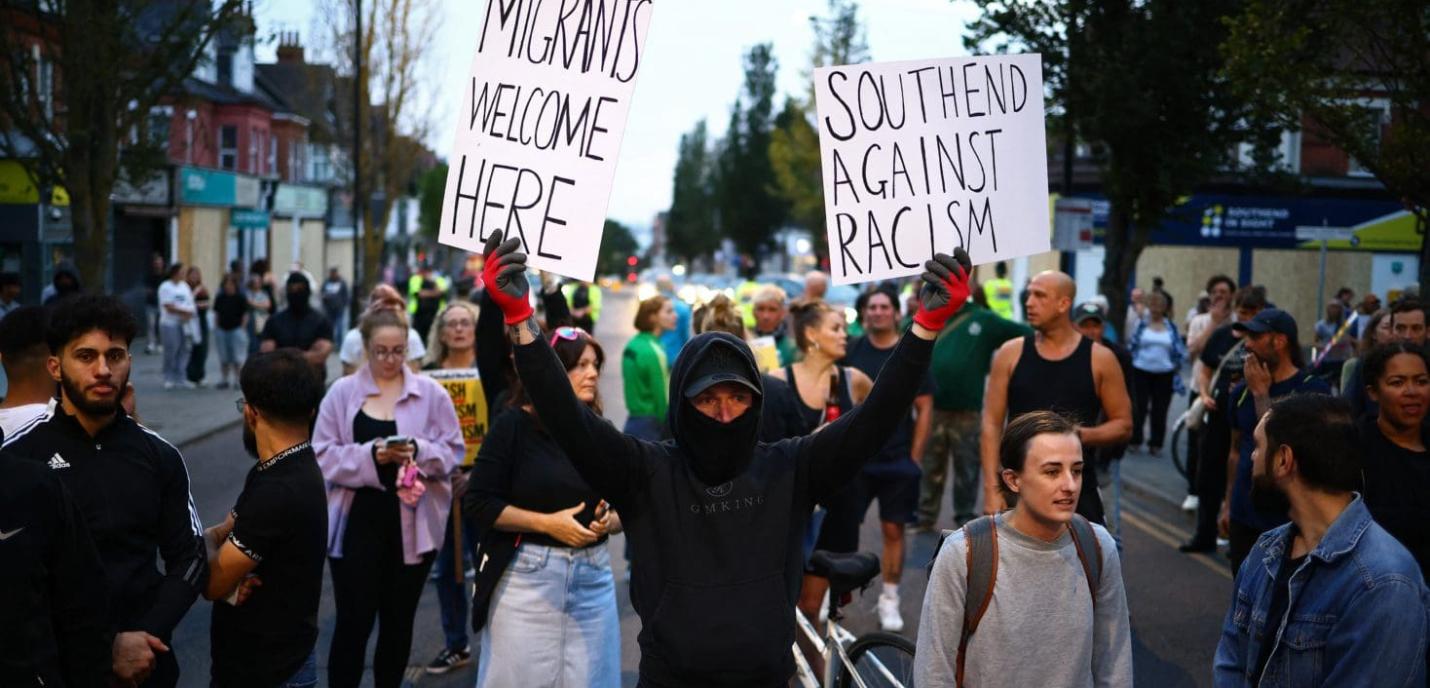 Manifestation dans une rue avec un groupe de personnes. Au centre, une personne masquée vêtue de noir tient deux pancartes en l'air indiquant : "Migrants welcome here" et "Southend against racism". D'autres manifestants sont visibles en arrière-plan