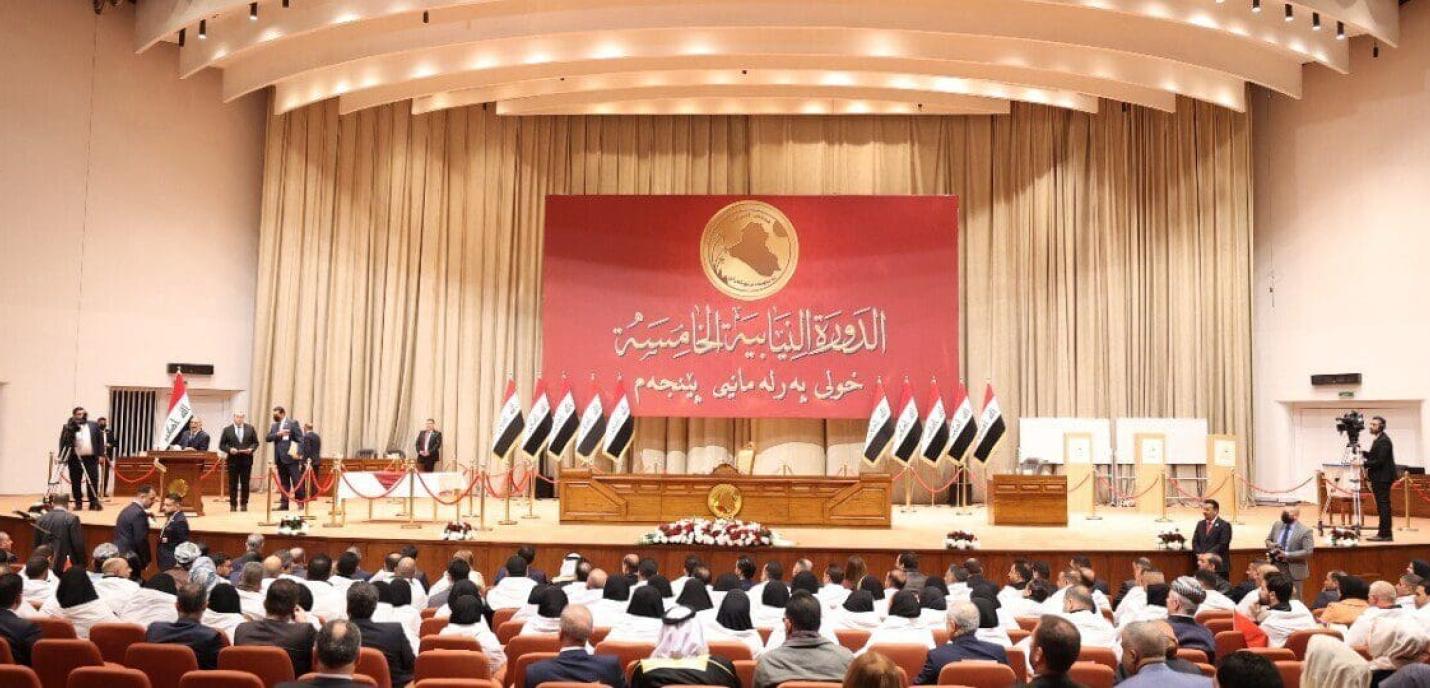 « Vue d’ensemble d’une séance parlementaire dans un grand hémicycle en Irak. Des parlementaires sont assis face à une estrade ornée de multiples drapeaux irakiens et d’un grand panneau rouge portant des inscriptions en arabe et un emblème doré représentant la carte du pays. Plusieurs responsables se tiennent debout sur la scène, tandis que des caméras et agents de sécurité sont présents dans la salle. 