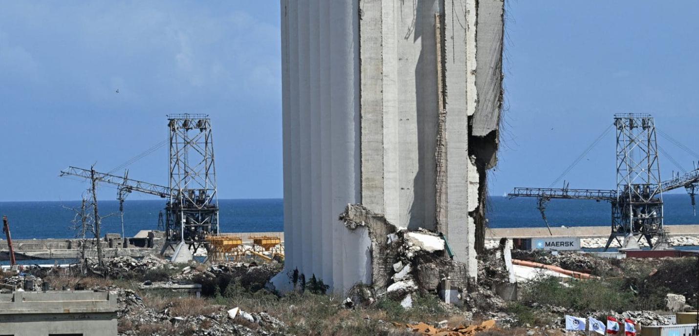 Vue sur un silo gravement endommagé à proximité du port, avec des gravats et des structures métalliques en arrière-plan. À l'horizon, on aperçoit la mer Méditerranée sous un ciel bleu. Des drapeaux libanais sont visibles dans le coin inférieur droit.