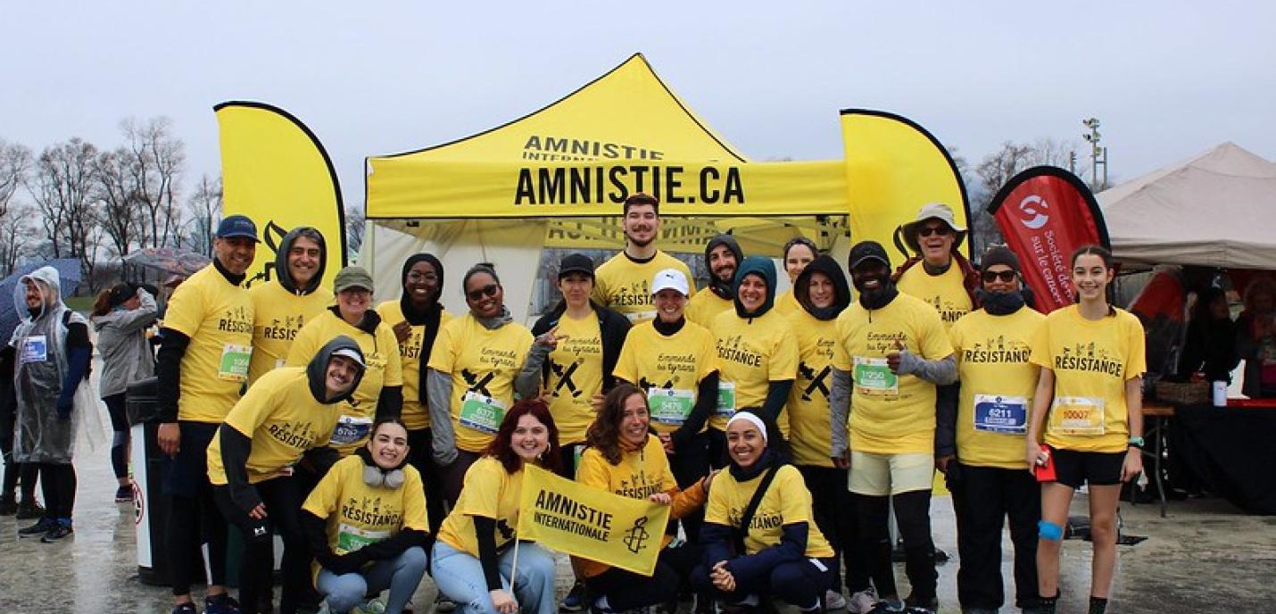 Un groupe de personnes souriantes posent ensemble devant une tente jaune d’Amnistie internationale sur laquelle on peut lire Amnistie.ca. Tous portent des t-shirts jaunes avec l’inscription “Résistance” et des dossards de course. Une femme au premier plan tient un petit drapeau d’Amnistie internationale. La scène se déroule lors d’un événement sportif extérieur, par temps pluvieux.
