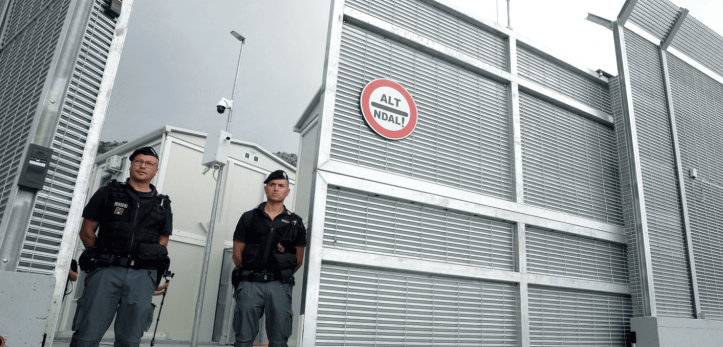« Deux agents de sécurité en uniforme noir se tiennent debout devant une haute clôture métallique munie de caméras de surveillance. Un panneau circulaire rouge et blanc portant l'inscription "ALT NDAL!" est visible sur la grille. L'arrière-plan montre des structures modulaires blanches sous un ciel gris. 