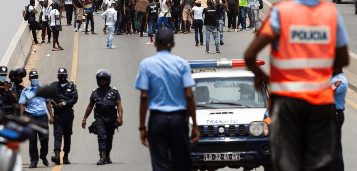 « Manifestation sur une route, avec un groupe de personnes rassemblées en arrière-plan. Au premier plan, plusieurs policiers, certains portant des casques et des gilets, encadrent la scène, tandis qu'une voiture de police marquée "Trânsito" est visible. Un policier en gilet fluorescent observe la situation