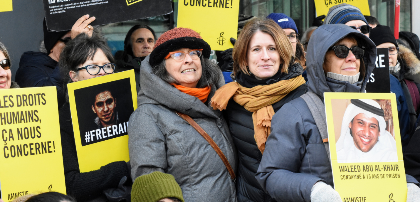 Un groupe de militants d’Amnistie internationale participe à une manifestation en plein air par temps froid. Les personnes portent des pancartes jaunes et noires avec des slogans sur les droits humains, dont « Les droits humains, ça nous concerne ! », « #FreeRaif » et une photo de Waleed Abu al-Khair avec la mention « condamné à 15 ans de prison ». Les participants sont emmitouflés dans des manteaux et bonnets d’hiver, certains sourient à la caméra.