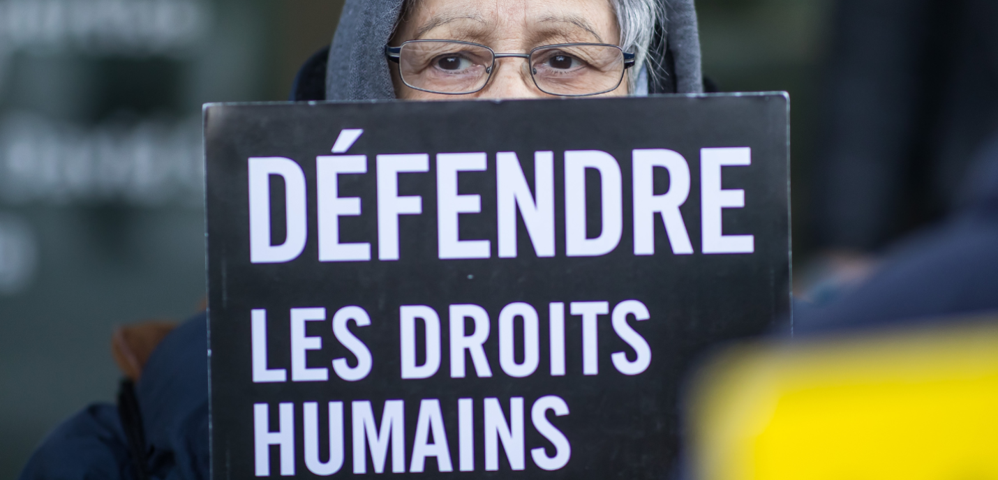 Une personne âgée portant des lunettes et une capuche grise tient une pancarte noire sur laquelle est écrit en blanc « DÉFENDRE LES DROITS HUMAINS ». Son regard sérieux dépasse au-dessus de la pancarte, soulignant le message de défense des droits humains.