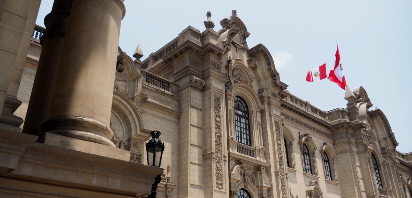 Un bâtiment gouvernemental imposant de style colonial, avec une façade en pierre richement décorée et de grandes colonnes. Deux drapeaux du Pérou flottent au sommet du bâtiment, sur fond de ciel bleu clair.