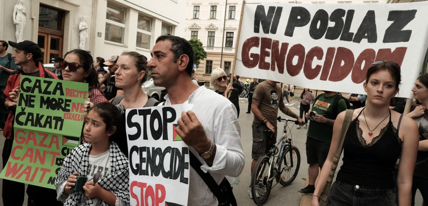 Un groupe de personnes participe à une manifestation dans la rue. Elles tiennent des pancartes portant des slogans contre le génocide et en soutien à Gaza. On peut lire « STOP GENOCIDE », « GAZA CAN'T WAIT » et « NI POSLA Z GENOCIDOM ». Les manifestants, dont des adultes et un enfant portant un keffieh, affichent des expressions sérieuses. L’ambiance est calme mais déterminée.