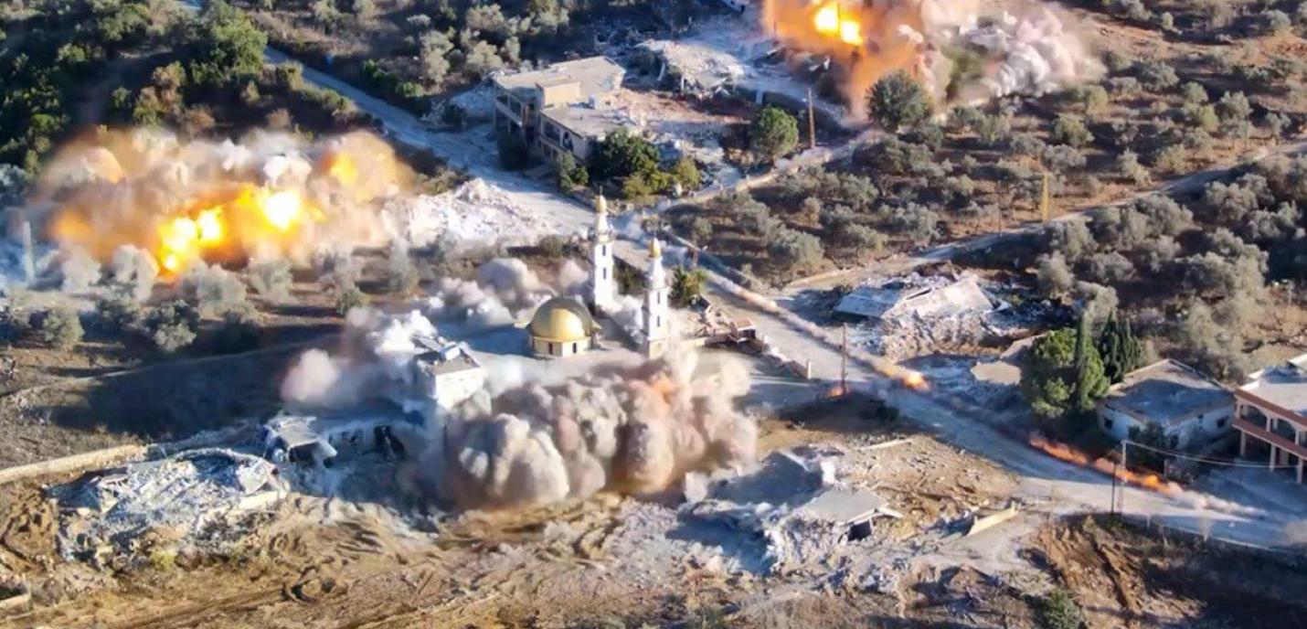Vue aérienne d’un village touché par des bombardements, avec plusieurs explosions visibles et un épais nuage de fumée s’élevant autour d’une mosquée à coupole dorée, au milieu de bâtiments détruits et de zones rurales.