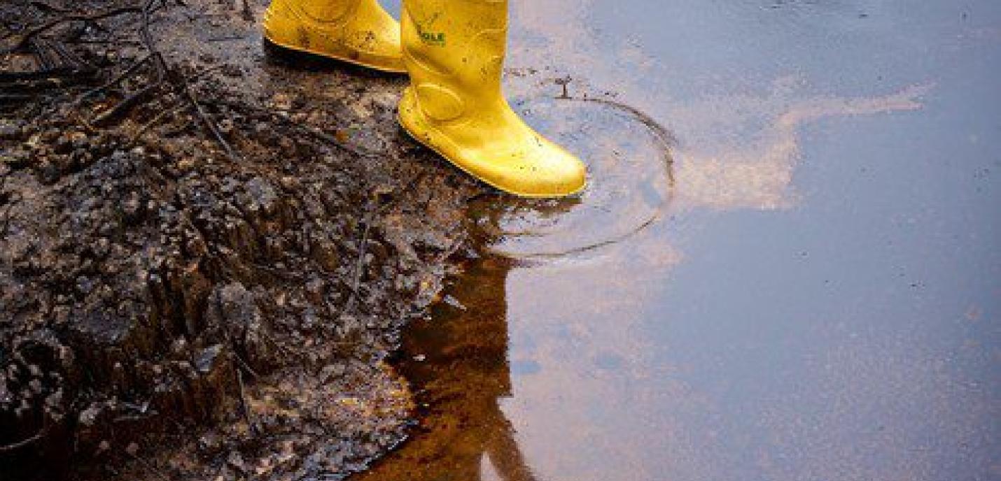 Une personne portant des bottes jaunes en caoutchouc se tient sur un sol boueux et souillé par une nappe de pétrole, dans une zone polluée par des hydrocarbures.