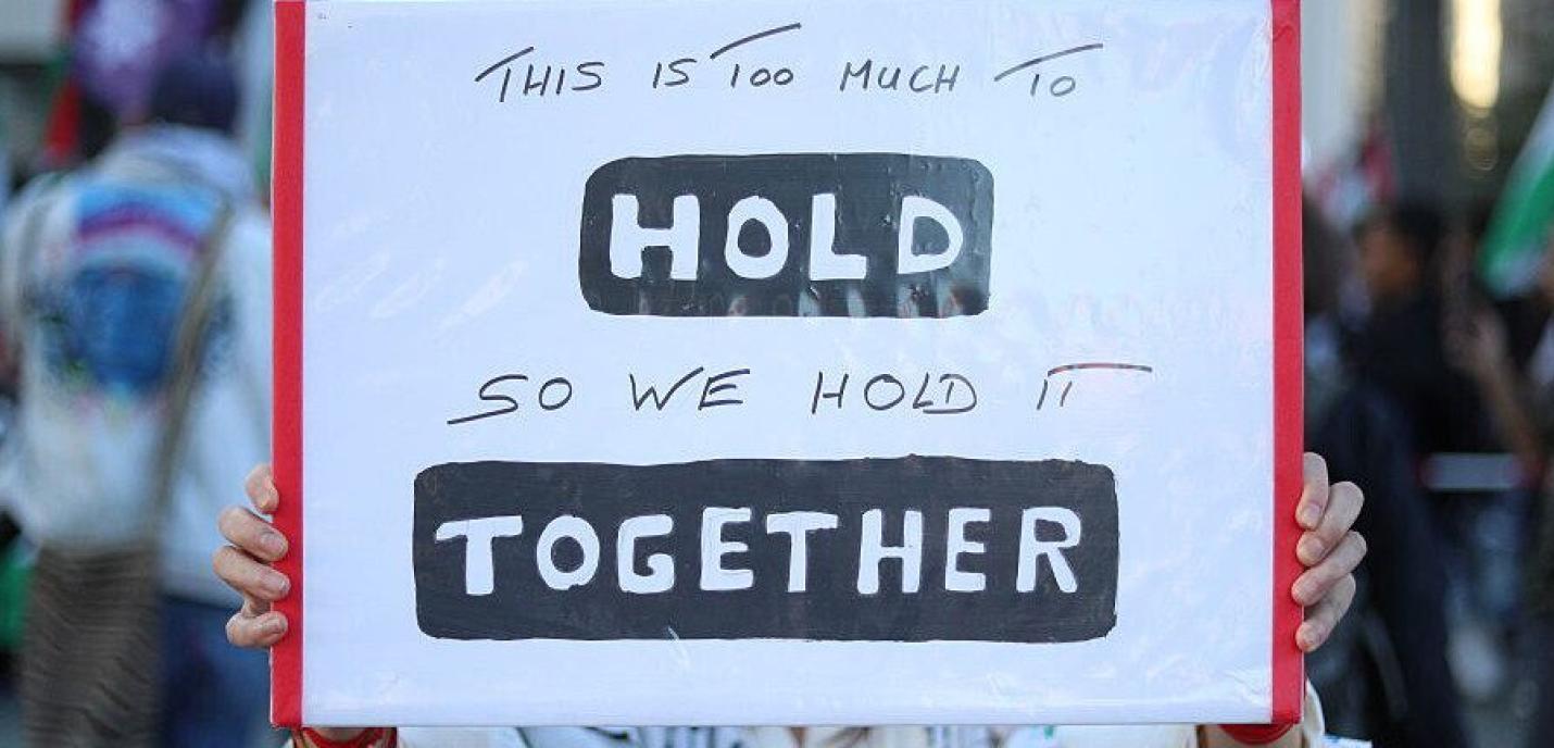 Une pancarte tenue par une personne lors d’une manifestation. Le panneau blanc, bordé de rouge, porte un message manuscrit en anglais : « This is too much to HOLD so we hold it TOGETHER ». Les mots « HOLD » et « TOGETHER » sont mis en évidence en lettres majuscules blanches sur fond noir. En arrière-plan flou, d’autres manifestants et drapeaux palestiniens apparaissent.