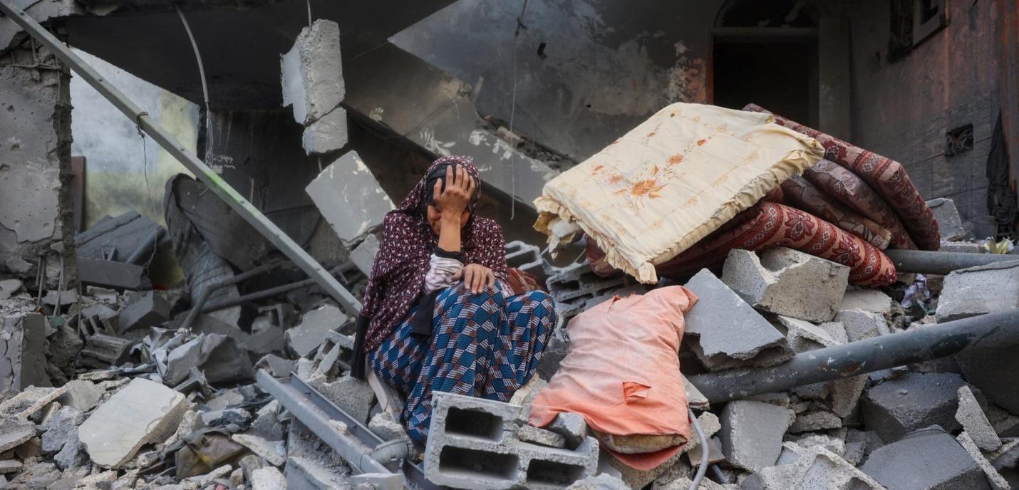 Une femme assise au milieu des décombres d’un bâtiment détruit, la tête dans la main en signe de détresse. Autour d’elle, des blocs de béton brisés, des poutres métalliques tordues et quelques matelas recouverts de poussière témoignent de la destruction causée par le bombardement.