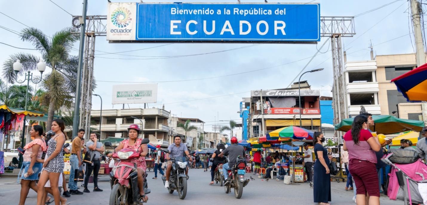 Plusieurs personnes circulent à scooter devant un panneau "Bienvenue en Équateur"