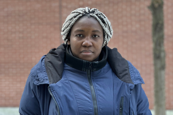 Photo portrait de Bénédicte-Carole, un travailleuse Camerounaise. Elle est devant un mur en brique rouge avec une veste d'hiver bleue.