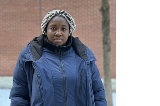 Photo portrait de Bénédicte-Carole, un travailleuse Camerounaise. Elle est devant un mur en brique rouge avec une veste d'hiver bleue.