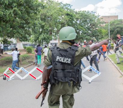 Un homme habillé en uniforme de police se tient à l'avant plan de l'image, dos à la caméra. Des manifestant·e·s sont dispersé·e·s à l'arrière-plan.