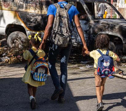 Un père emmène ses deux enfants à l'école, tenant leur main. Devant eux, une voiture calcinée bloque la rue.